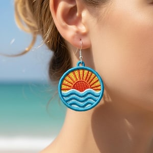 May include: Embroidered round earrings with a sunset design. The earrings feature a yellow sun, orange rays, and blue waves, with a blue border and silver hook. These earrings are a beach-themed accessory.