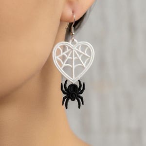 May include: A dangling earring featuring a white embroidered heart shape with an internal spider web pattern. A small, black embroidered spider is attached to the bottom of the heart. The earring hangs from a silver-toned metal hook wire.