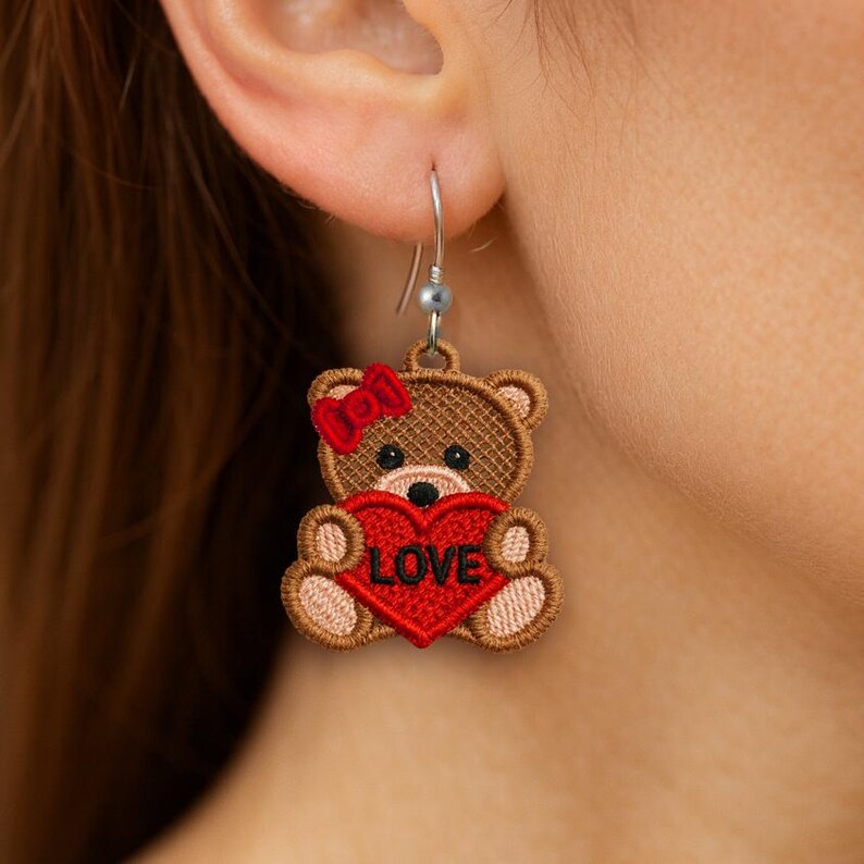 May include: A single teddy bear earring with a red heart that says "LOVE". The brown bear has a red bow and is embroidered. The earring is hanging from a silver hook.