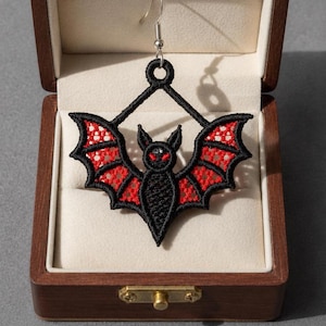May include: A black and red embroidered bat earring featuring a detailed stitched design with a central bat figure, red wing accents, and a silver-toned metal hook. The earring is displayed inside a rectangular wooden box with a cream-colored interior.