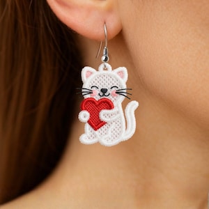 May include: White embroidered cat-shaped earring holding a red heart. The cat has black whiskers and a smiling face. The earring is suspended from a silver-colored hook.