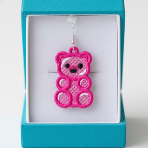 Könnte beinhalten: Ein pinkfarbener, gestickter Teddybär-Ohrring mit schwarzen Augen und weißen Akzenten, präsentiert in einer türkisfarbenen Schmuckschatulle. Der Ohrring ist ein detailliertes, durchbrochenes Design, das die Form des Bären hervorhebt. Die Schatulle bietet einen kontrastierenden Hintergrund.