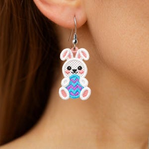 Può includere: Un orecchino ricamato a forma di coniglietto pasquale bianco con dettagli rosa e blu. Il coniglietto tiene in mano un uovo di Pasqua blu e viola. L'orecchino ha un gancio argentato.
