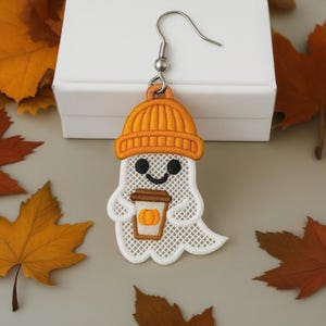 Puede incluir: Un solo pendiente con tem&aacute;tica de Halloween con un fantasma blanco que lleva un gorro naranja y sostiene una taza de caf&eacute; con un dise&ntilde;o de calabaza. El pendiente est&aacute; sobre un fondo de hojas de oto&ntilde;o y una caja blanca.