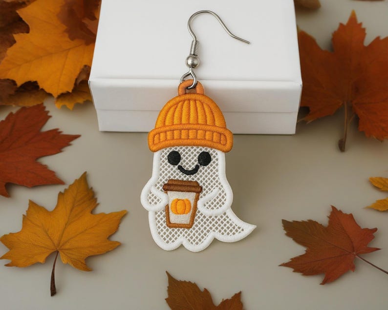 Puede incluir: Un solo pendiente con tem&aacute;tica de Halloween con un fantasma blanco que lleva un gorro naranja y sostiene una taza de caf&eacute; con un dise&ntilde;o de calabaza. El pendiente est&aacute; sobre un fondo de hojas de oto&ntilde;o y una caja blanca.