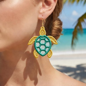 May include: A gold-colored, embroidered sea turtle earring with a teal and white center, hanging from a gold hook. The earring is worn by a person with a beach scene in the background.