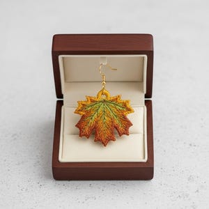 Puede incluir: Un pendiente individual de hoja de arce bordada en una caja de joyería de madera. La hoja presenta tonos de verde, amarillo y naranja, con un gancho dorado. La caja está abierta, revelando un interior color crema.