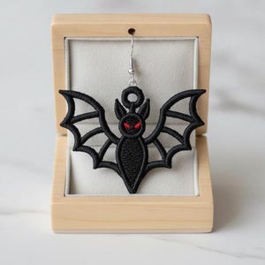 May include: A black embroidered bat earring with red eyes and a silver-toned metal hook, presented inside a light-colored wooden box with a soft, light gray interior lining. The bat features detailed stitching and a symmetrical wing design.