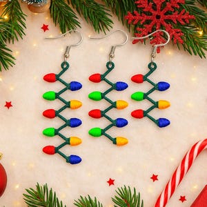 May include: Three pairs of Christmas light bulb earrings. Each earring features a green zig-zag design with red, orange, green, and blue bulbs. The earrings are set against a festive background with ornaments and greenery.