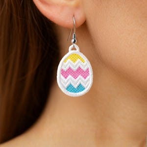 May include: Embroidered Easter egg earring with a white border and a colorful zig-zag pattern in yellow, pink, and blue. The earring is suspended from a silver-colored hook. The earring is worn on a person's ear.