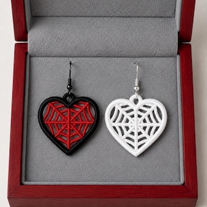 May include: A pair of heart-shaped drop earrings featuring a spiderweb design. One earring has a black frame with red web detailing and a black hook, while the other has a white frame with white web detailing and a silver-toned metal hook.
