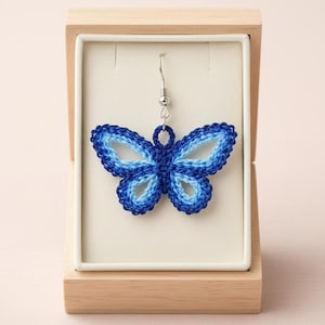 May include: A blue butterfly earring displayed in a wooden box. The earring is made of a woven, gradient blue material, with shades ranging from light to dark blue. The earring is suspended from a silver-colored hook.