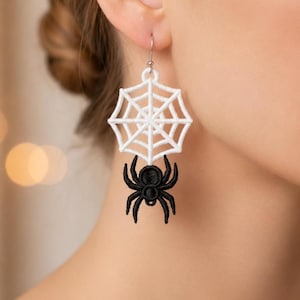 Puede incluir: Un pendiente colgante que presenta una forma de telaraña bordada en blanco suspendida de un gancho plateado, con una pequeña araña negra bordada colgando del centro inferior de la red, creando un diseño monocromático de alto contraste.