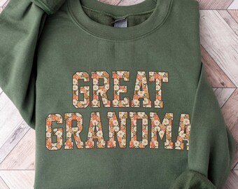 May include: A green sweatshirt with the text "GREAT GRANDMA" in white letters outlined in orange. The letters are filled with a floral pattern of orange, white, and brown.