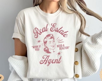 May include: A white t-shirt with a pink graphic that says "Real Estate Agent" with a vintage-style illustration of a woman and the text "Wink if you need a realtor".