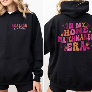 May include: Black hooded sweatshirt with the text "REALTOR Kimberly" on the front and "IN MY HOME MATCHMAKER ERA" with pink and orange lettering and floral accents on the back.