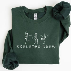 May include: A dark green sweatshirt with the text "Skeleton Crew" printed in white. Three white skeleton figures are dancing in front of the text.