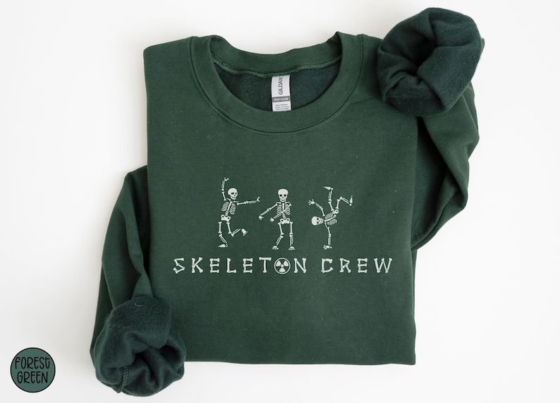 May include: A dark green sweatshirt with the text "Skeleton Crew" printed in white. Three white skeleton figures are dancing in front of the text.