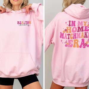 May include: A light pink hoodie with the text "REALTOR Kimberly" on the front and "IN MY HOME MATCHMAKER ERA" on the back. The text is in a retro font with colorful accents.