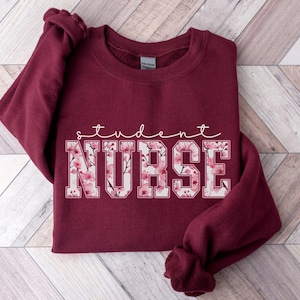 Może przedstawiać: Bordowa bluza z napisem "student nurse" w białych literach obwiedzionych czarną ramką. Litery są wypełnione r&oacute;żowym wzorem kwiatowym.