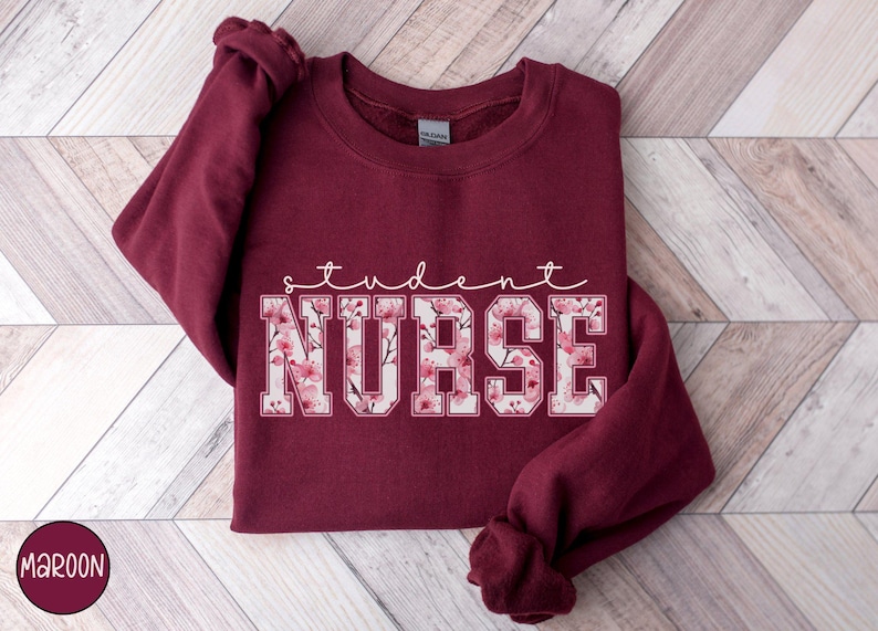 Może przedstawiać: Bordowa bluza z napisem "student nurse" w białych literach obwiedzionych czarną ramką. Litery są wypełnione r&oacute;żowym wzorem kwiatowym.