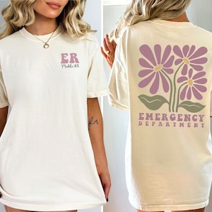 May include: Two ivory t-shirts with different designs. The front of the first shirt has the letters "ER" and the text "Middle RN". The back of the second shirt has a design of three purple flowers with green leaves and the text "Emergency Department".