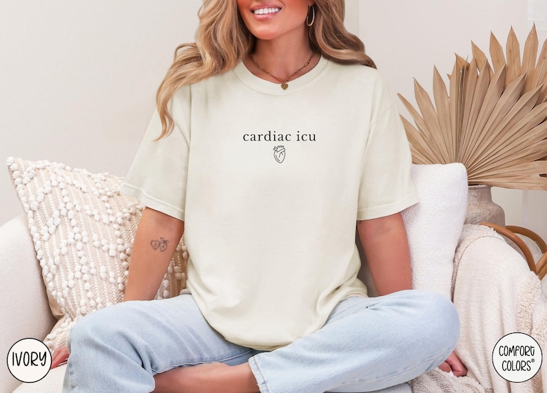 Pu&ograve; includere: Una donna indossa una maglietta bianca con il testo "cardiac icu" e una piccola grafica a forma di cuore. &Egrave; seduta su un divano con un cuscino bianco e una foglia di palma marrone.