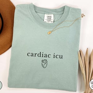 Pu&ograve; includere: Una maglietta verde chiaro con il testo "cardiac icu" e una piccola grafica a forma di cuore stampata sul davanti.