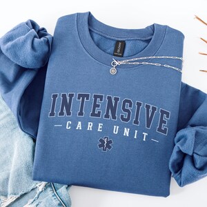 May include: A blue sweatshirt with the text "INTENSIVE CARE UNIT" in white letters with a black outline. A small medical symbol is below the text.