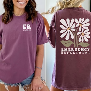 May include: A purple t-shirt with the text "ER" and "Middle TN" on the front and a design of three white daisies with green leaves and the text "EMERGENCY DEPARTMENT" on the back.