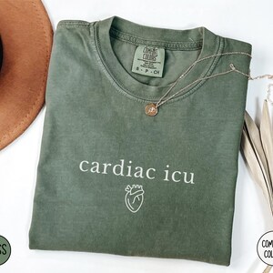 Pu&ograve; includere: Una maglietta verde con il testo "cardiac icu" e una piccola grafica a forma di cuore. La maglietta &egrave; realizzata da Comfort Colors.