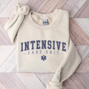 May include: A beige sweatshirt with the text "INTENSIVE CARE UNIT" in blue lettering and a blue star of life symbol.