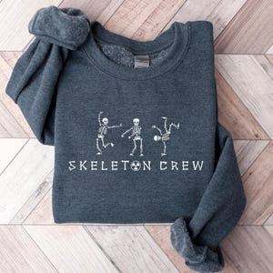 May include: Dark heather grey sweatshirt with a white graphic of three skeletons dancing and the text "Skeleton Crew".