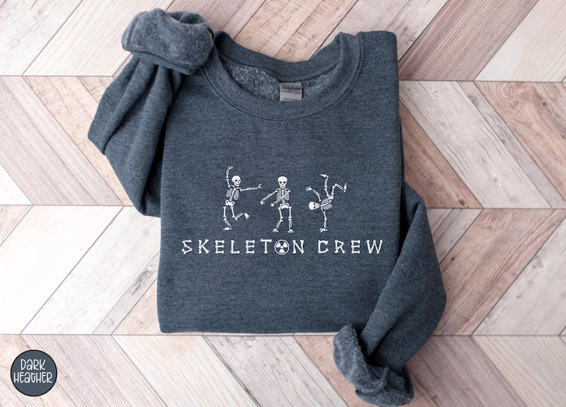 May include: Dark heather grey sweatshirt with a white graphic of three skeletons dancing and the text "Skeleton Crew".