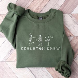 May include: A green sweatshirt with a white graphic of three skeletons dancing and the text "Skeleton Crew".