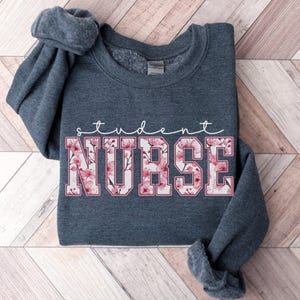 Może przedstawiać: Szara bluza w kolorze ciemnego melanżu z napisem "student nurse" białymi literami z r&oacute;żowym wzorem kwiatowym.