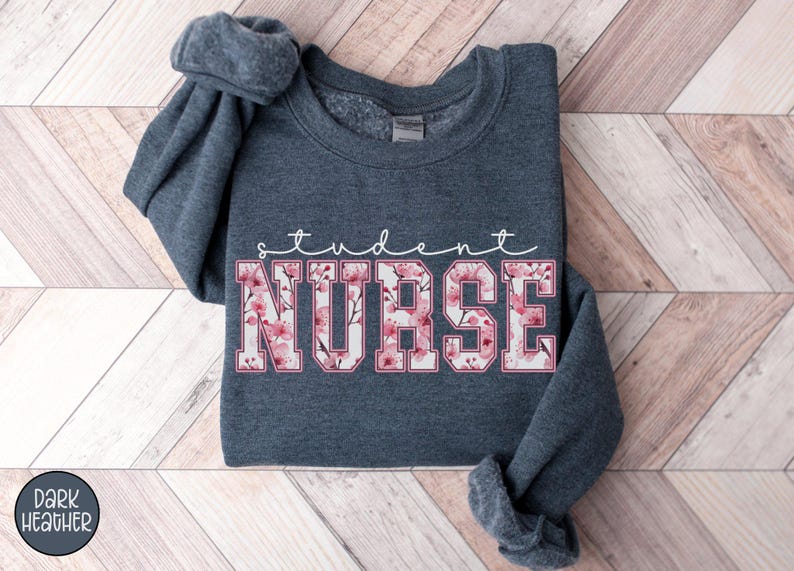 Może przedstawiać: Szara bluza w kolorze ciemnego melanżu z napisem "student nurse" białymi literami z r&oacute;żowym wzorem kwiatowym.