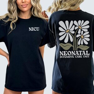 Może przedstawiać: Czarny T-shirt z napisem "NICU" z przodu i wzorem białych stokrotek z zielonymi łodygami i liśćmi z tyłu. Napis "Neonatal Intensive Care Unit" znajduje się pod stokrotkami.