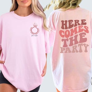 May include: Two pink t-shirts with different designs. The first shirt has a ring with a diamond and the text "Here comes the party Jennifer". The second shirt has the text "Here comes the party" in a retro font.