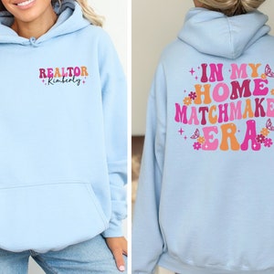 May include: Light blue hoodie with the text "REALTOR +Kimberly+" on the front and "IN MY HOME MATCHMAKER ERA" with pink and orange lettering and flowers on the back.