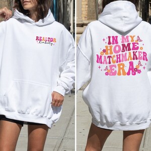 May include: White hooded sweatshirt with the text "REALTOR Kimberly" on the left side and "IN MY HOME MATCHMAKER ERA" with pink and orange lettering and butterfly graphics on the right side.