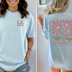 May include: Light blue t-shirt with the text "ER" and "Med Tech RN" on the front. The back of the shirt has a pink floral design with the text "Emergency Department".
