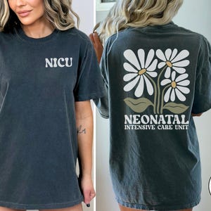 Może przedstawiać: Szary t-shirt z napisem "NICU" z przodu i "NEONATAL INTENSIVE CARE UNIT" z kwiatowym wzorem z tyłu. Koszulka wykonana jest z miękkiego, wygodnego materiału.