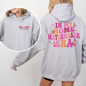 May include: A gray hooded sweatshirt with the text "REALTOR Kimberly" on the front and "IN MY HOME MATCHMAKER ERA" on the back. The text is in a retro font with pink, orange, and yellow colors. The back also features pink flowers and butterflies.