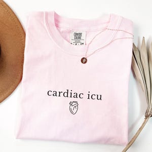 Pu&ograve; includere: Una maglietta rosa con il testo "cardiac icu" e una piccola grafica a forma di cuore. La maglietta &egrave; realizzata da Comfort Colors.