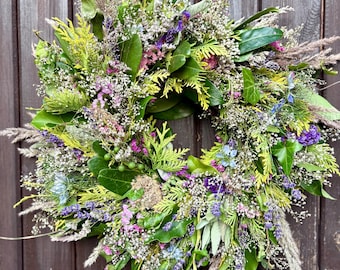 Könnte beinhalten: Ein kreisförmiger Kranz aus verschiedenen getrockneten Blumen und Grünpflanzen, darunter Lavendel, Weiß und grünes Laub. Der Kranz hängt vor einer dunkelbraunen Holztür und zeigt eine natürliche, rustikale Ästhetik.