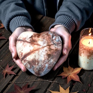 Op de afbeelding: Een hartvormige steen met bruine en crèmekleurige adering wordt in open handen gehouden. De steen ligt op een houten oppervlak met herfstbladeren en een brandende kaars in een glas.