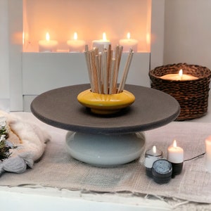 Op de afbeelding: Een zwart-witte keramische sokkel met een gele keramische kom erop. De kom bevat een bos houten stokjes. De sokkel staat op een beige oppervlak met kaarsen en een rieten mand.