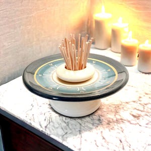 Op de afbeelding: Een decoratieve tandenstokerhouder van keramiek. De houder heeft een crèmekleurige basis en een bijpassende ring om de tandenstokers in te plaatsen. De plaat heeft een turquoise en gouden ontwerp met een zwarte rand. Een bundel houten tandenstokers.
