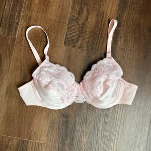 May include: A light pink underwire bra featuring a satin lower cup and a floral lace upper cup. The garment includes adjustable shoulder straps, a small center bow detail, and scalloped lace edges along the top of the cups and the center bridge.
